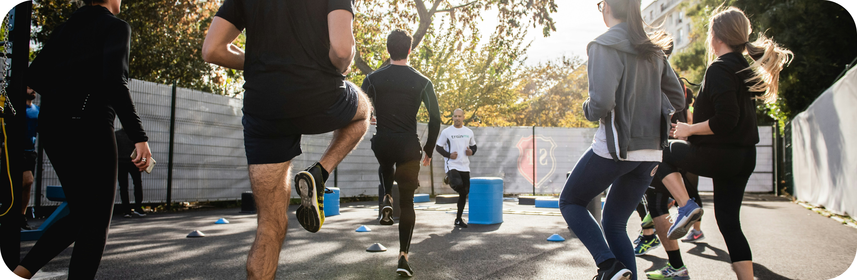 Group of people doing outdoor fitness training, performing high-knee running drills around cones in a park with a coach leading the session.