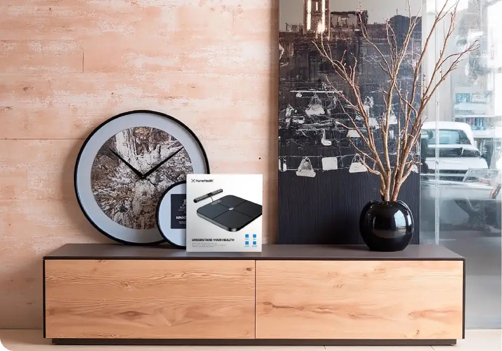 Modern home interior with wooden cabinet displaying Hume Body Pod scale packaging, decorative clock, and vase, showcasing product in a lifestyle setting.